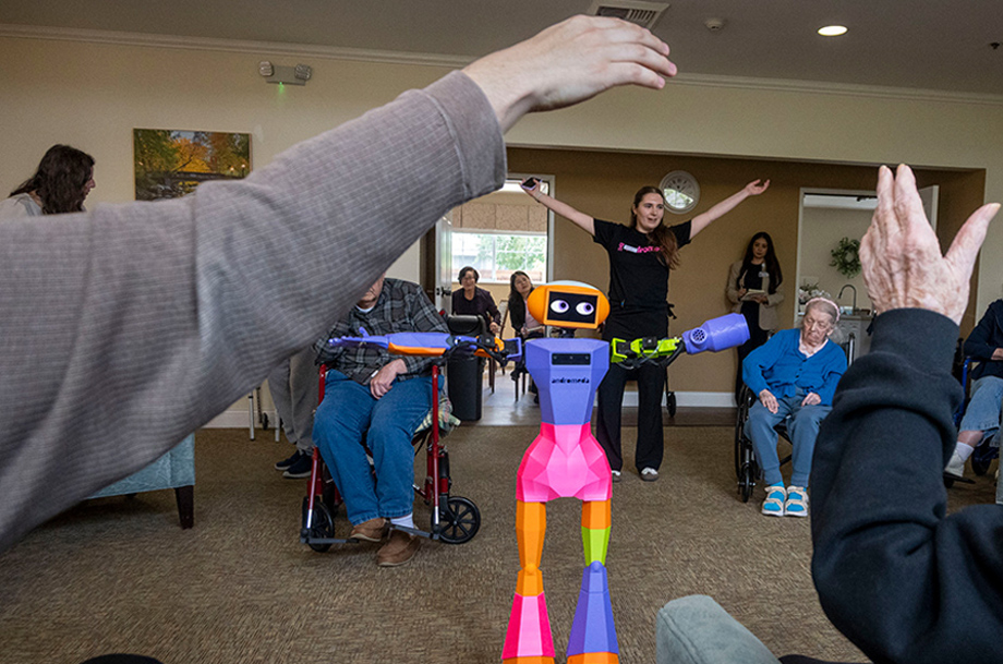 Group exercise in a senior living room, with residents raising arms while a colorful robot and instructor lead the activity.